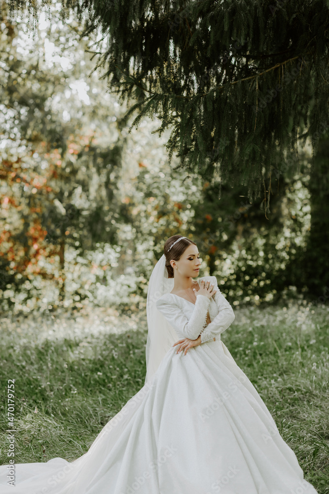 The bride in a beautiful white dress stands in front of a tree