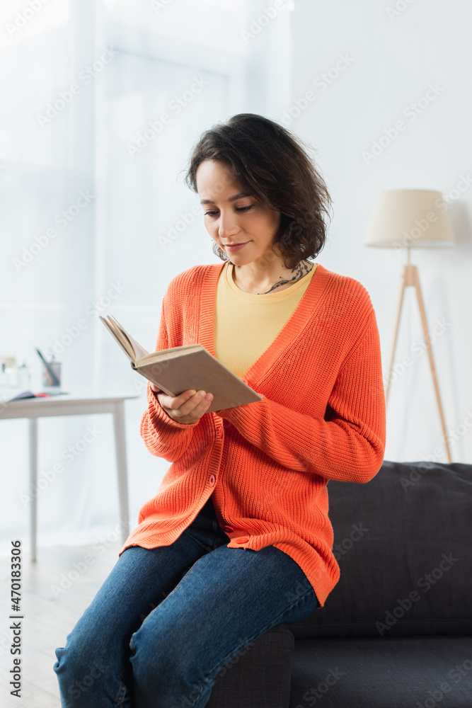 tattooed woman reading book while sitting on couch at home. tattooed woman reading book while sitting on couch at home.
