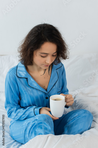 young woman in blue pajamas holding cup of coffee in bedroom.