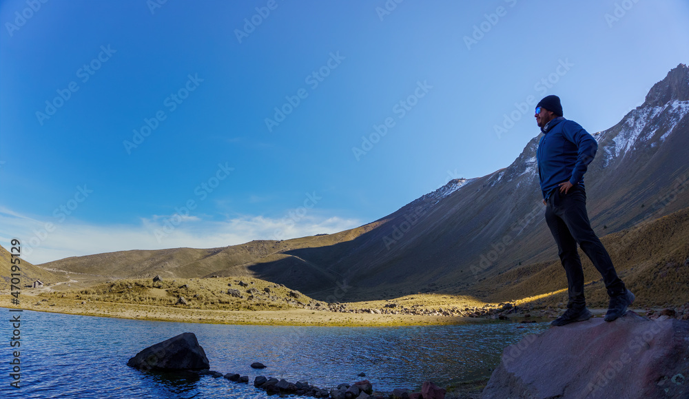 Young hiker wearing blue jacket standing by the mountain lake and looking at the distance