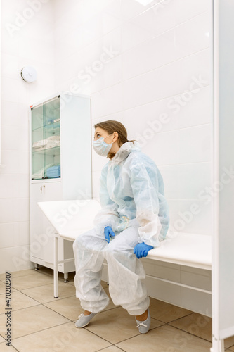 tired and sad female doctor in medical uniform  blue gloves  mask and glasses  sitting in a room for medical procedures