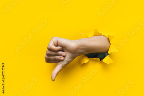 Hand showing a thumb down through ripped hole in bright yellow paper background. Concept of dislike and disapproval gesture.