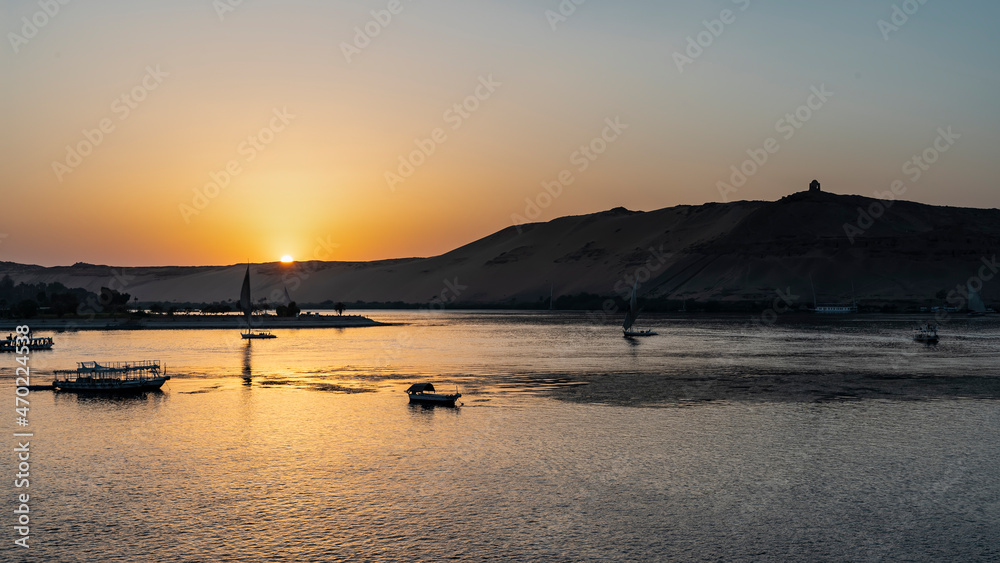 Fototapeta premium Sunset on the Nile. The sun hides behind the sand dunes. The sky is highlighted in orange. Several boats can be seen on the river. Glare on the water. Egypt