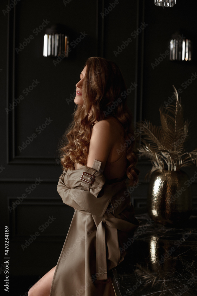 Sensual adorable model girl with curly hairstyle wearing trench coat poses in the luxurious interior