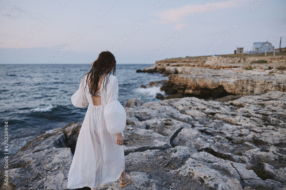 woman with wet hair in a white dress is walking along the rocky shore