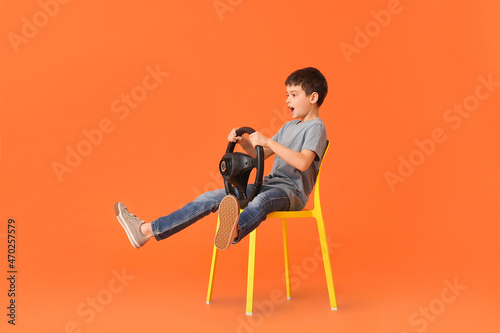Surprised little boy with steering wheel sitting in chair on color background