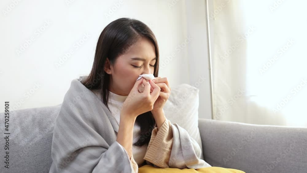 Young Sick Asian Woman Sitting under the Blanket Whiles Sneezing with Tissue on the Sofa Young Sick Asian Woman Sitting under the Blanket Whiles Sneezing with Tissue on the Sofa