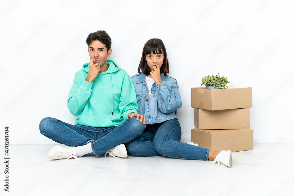 Young couple making a move while picking up a box full of things sitting on the floor isolated on white background having doubts while looking up Young couple making a move while picking up a box full of things sitting on the floor isolated on white background having doubts while looking up