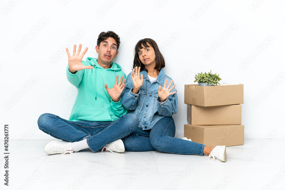 Young couple making a move while picking up a box full of things sitting on the floor isolated on white background is a little bit nervous and scared stretching hands to the front Young couple making a move while picking up a box full of things sitting on the floor isolated on white background is a little bit nervous and scared stretching hands to the front