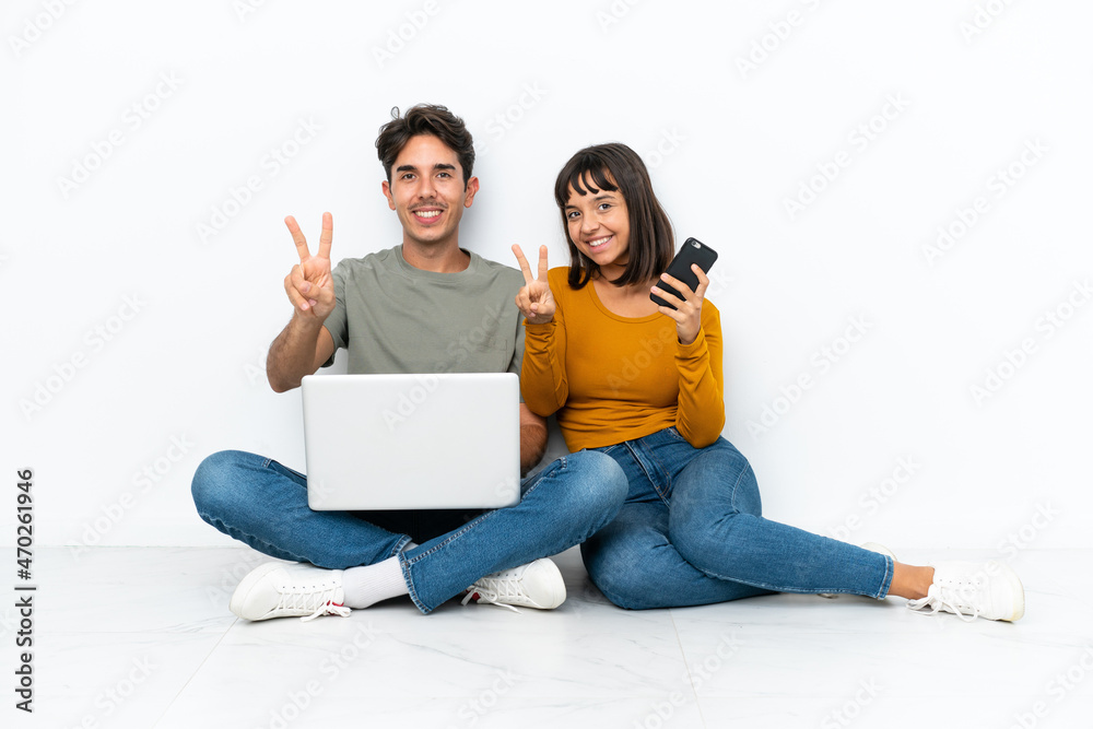 Young couple with a laptop and mobile sitting on the floor smiling and showing victory sign