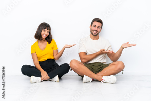 Young couple sitting on the floor isolated on white background pointing back and presenting a product