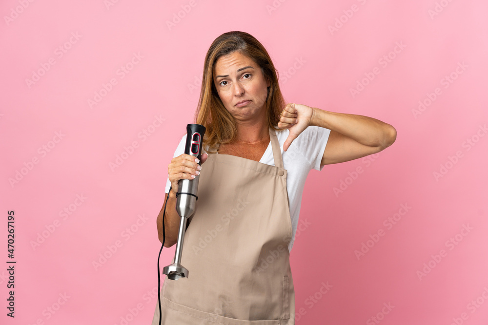 Middle age woman using hand blender isolated on pink background showing thumb down with negative expression Middle age woman using hand blender isolated on pink background showing thumb down with negative expression