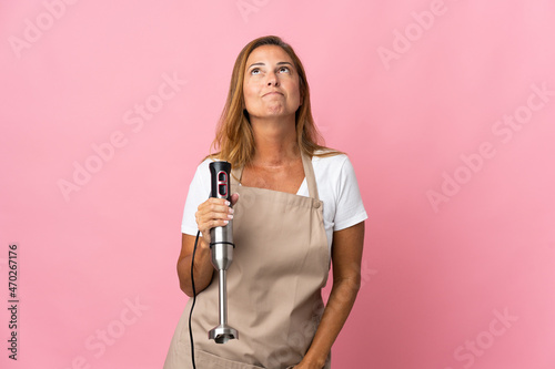 Middle age woman using hand blender isolated on pink background and looking up