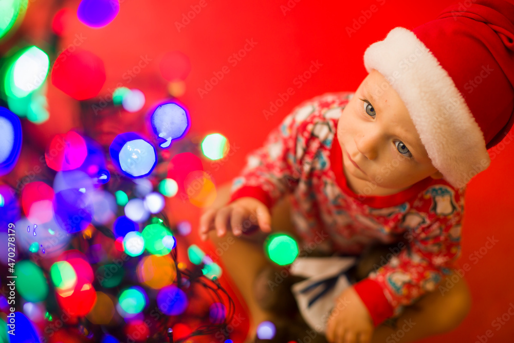 baby in santa claus hat on a red background looks at the colorful lights from garlands. baby in santa claus hat on a red background looks at the colorful lights from garlands.