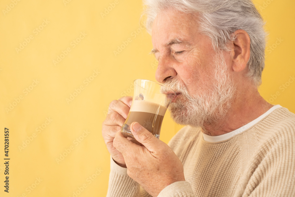Handsome bearded senior man drinking coffee and milk cappuccino from a glass beaker , standing on yellow background Handsome bearded senior man drinking coffee and milk cappuccino from a glass beaker , standing on yellow background
