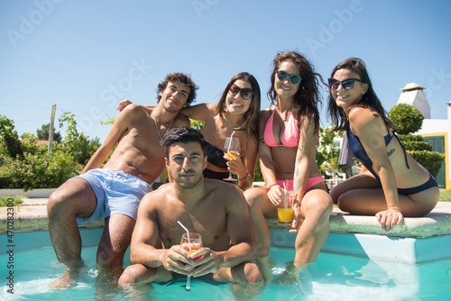 Contented young friends at poolside. Smiling young women and men in swimsuits resting in swimming pool, looking at camera. Leisure, friendship, party concept
