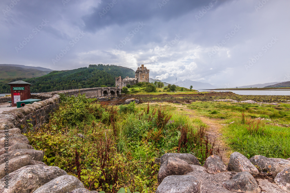 Landscape with Eilean Donan Castle in Scotland on an island. Historic stone footbridge leads to the castle. Hills and lake in the background. Stone wall with small wooden house and grass Landscape with Eilean Donan Castle in Scotland on an island. Historic stone footbridge leads to the castle. Hills and lake in the background. Stone wall with small wooden house and grass