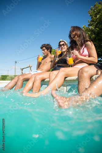 Happy young friends having fun near swimming pool. Men and women in swimsuits splashing water with feet, smiling, drinking cocktails. Leisure, friendship, party concept