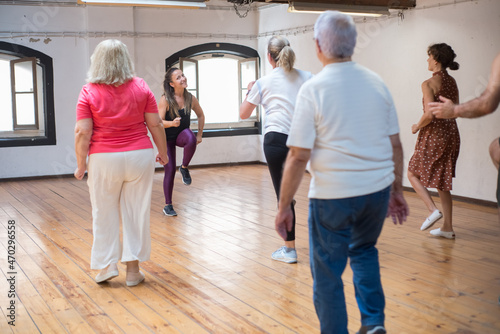 Funny young Caucasian teacher demonstrating dance steps. Cheerful female instructor with long fair hair showing dance moves to her senior group. Dance, hobby, healthy lifestyle concept