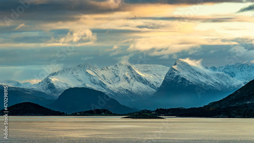 Wilde  schneebedeckte Berge in der Umgebung von   rnes  Norwegen  im orangen Morgenlicht bei Sturm mit Wolken. Schneefahnen wehen von den Bergspitzen herunter. Kreuzfahrt mit dem Postschiff 