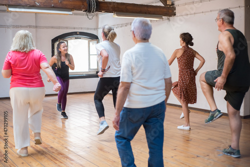 Hilarious young Caucasian instructor showing dance steps. Joyful female teacher with long hair in bright leggings demonstrating moves to her group of seniors. Dance, hobby, healthy lifestyle concept