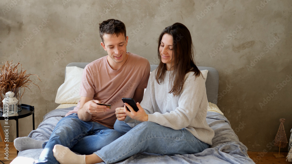 Young smiling husband and wife, paying for purchases and bills using phone and card, talking and laughing at home. Online shopping concept Young smiling husband and wife, paying for purchases and bills using phone and card, talking and laughing at home. Online shopping concept