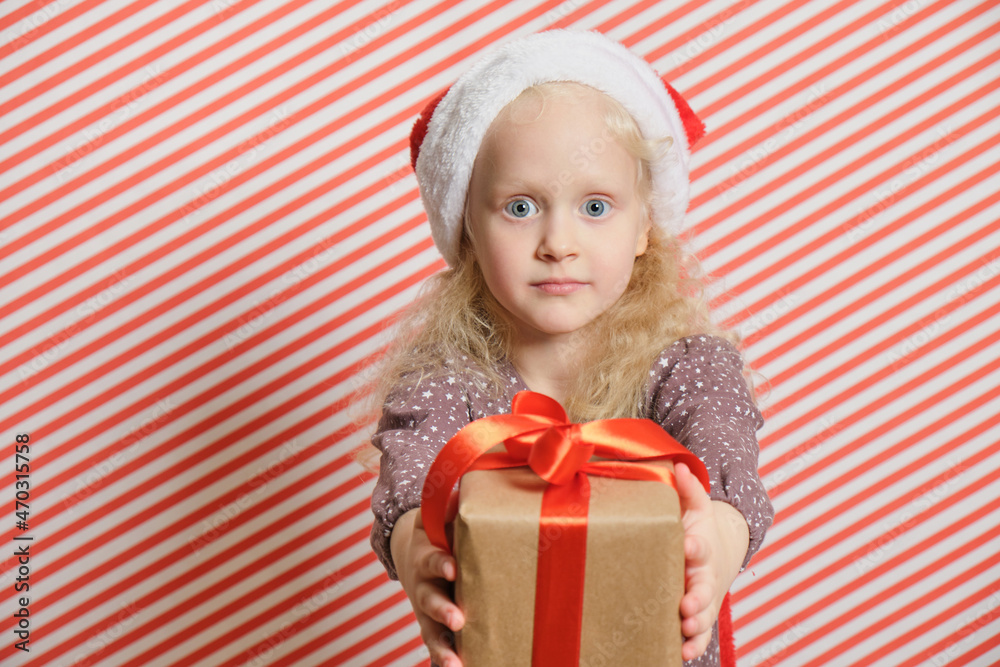 cheerful preschooler girl in a santa hat with a gift on a red striped background, boxing day, christmas gifts concept cheerful preschooler girl in a santa hat with a gift on a red striped background, boxing day, christmas gifts concept