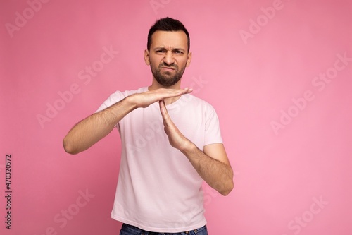 Photo of handsome unhappy angry dissatisfied brunet man in casual pink t-shirt isolated on pink background with copy space for text and showing time out and stop gesture with hands