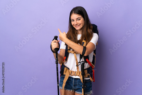 Young woman with backpack and trekking poles isolated on purple background pointing back