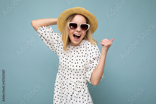 Photo of young positive happy attractive blonde woman with sincere emotions wearing summer dress, straw hat and sunglasses isolated over blue background with copy space and celebrating victory photo