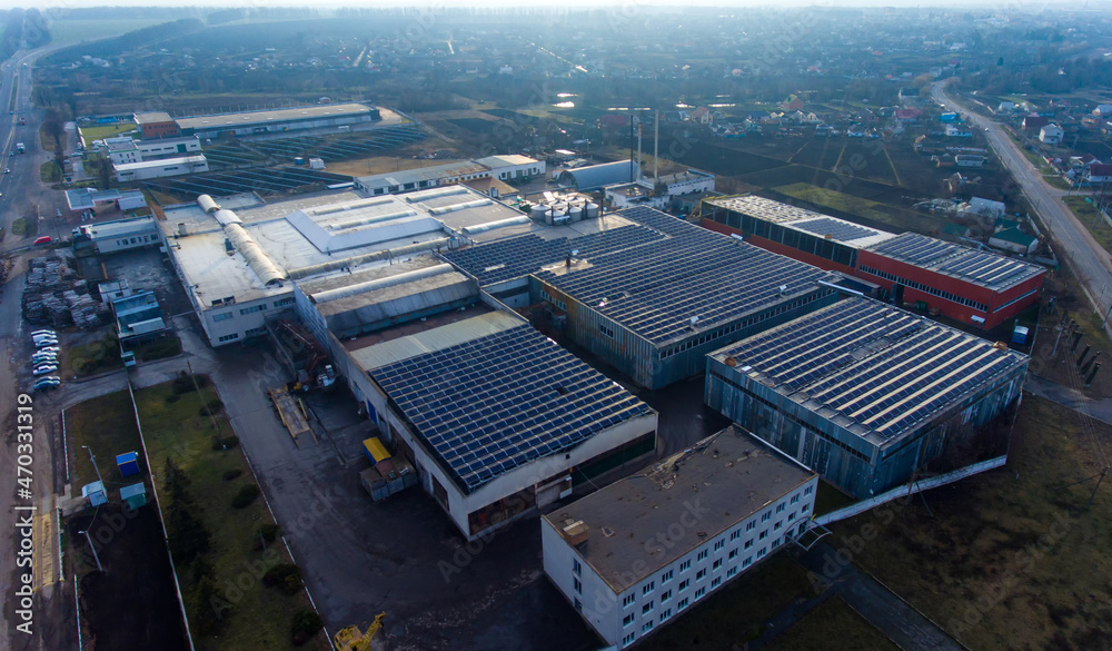 Numerous solar panels mounted on the industrial building. Applying spacious roof area for producing clean energy. View of the rural area at the background.