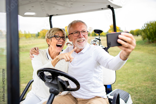 Smiling senior couple having fun at golf course and taking a selfie photo in golf car before training.