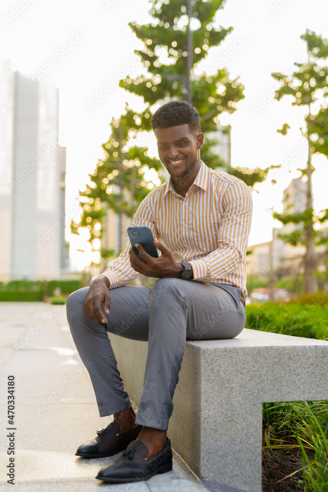 Obraz premium Full length portrait of young handsome African businessman sitting outdoors in city using mobile phone while smiling