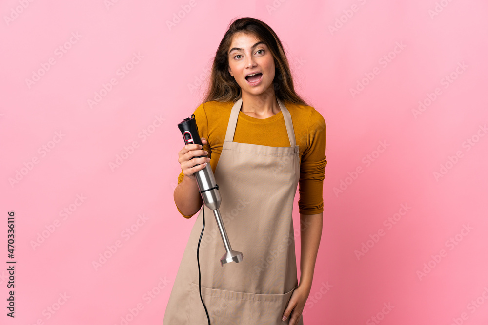 Young chef woman using hand blender isolated on pink background with surprise facial expression