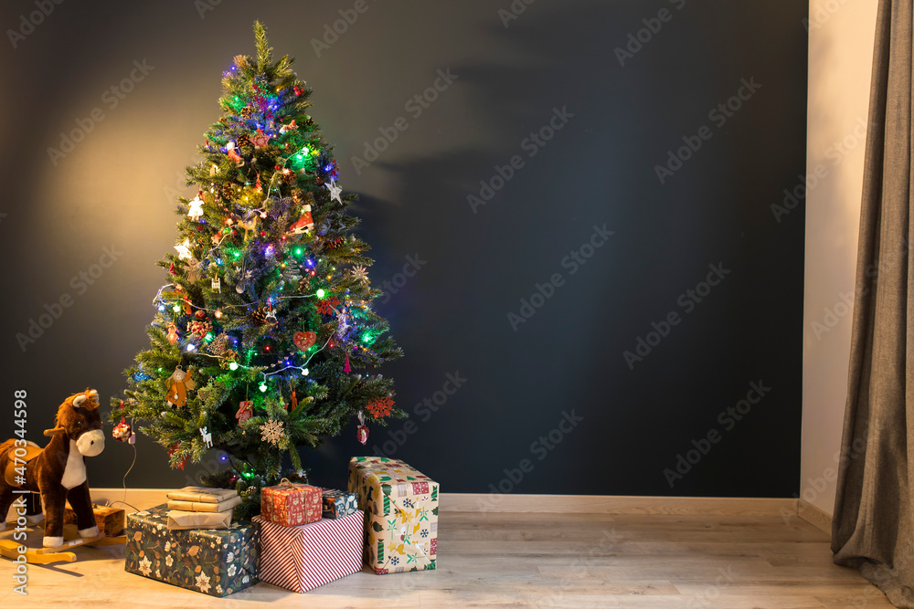 A decorated Christmas tree with a garland and wooden toys stands in the room on a dark blue background. Under the tree there are boxes with gifts and a rocking horse