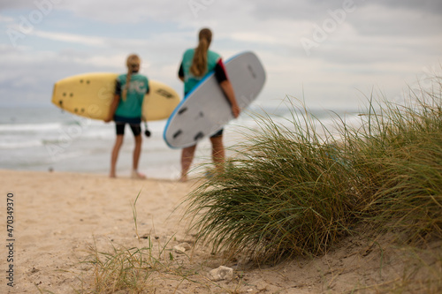 Bild auf Leinwand macro of grass in the foreground, blurry woman and girl with their surf boards o