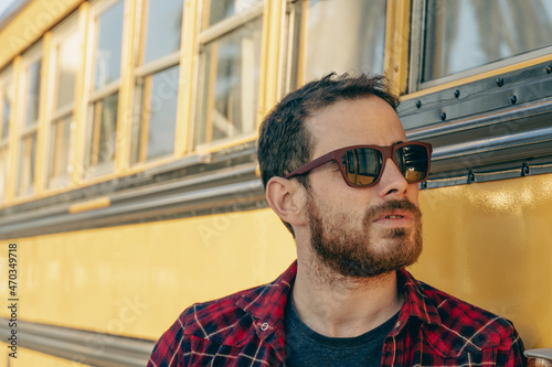 Young man in red plaid shirt and retro style with sunglasses observing an old yellow bus
