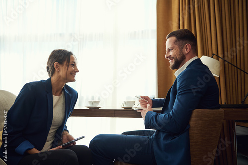 Handsome Caucasian man and attractive woman, business partners, investment advisors dressed in business suits sitting on armchair and discussing business matters in a hotel room during a business trip
