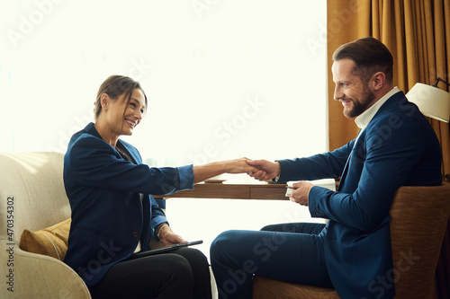 Fototapeta Naklejka Na Ścianę i Meble -  Attractive woman in a business suit smiles at her business partner while shaking hands, making agreement and closing deal at a meeting. Business negotiations in a hotel room during business trip