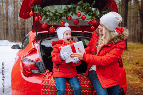 Happy family of blonde mom and little girl open Christmas gift box sitting in christmas decorated car in winter forest. Togetherness, holidays, family