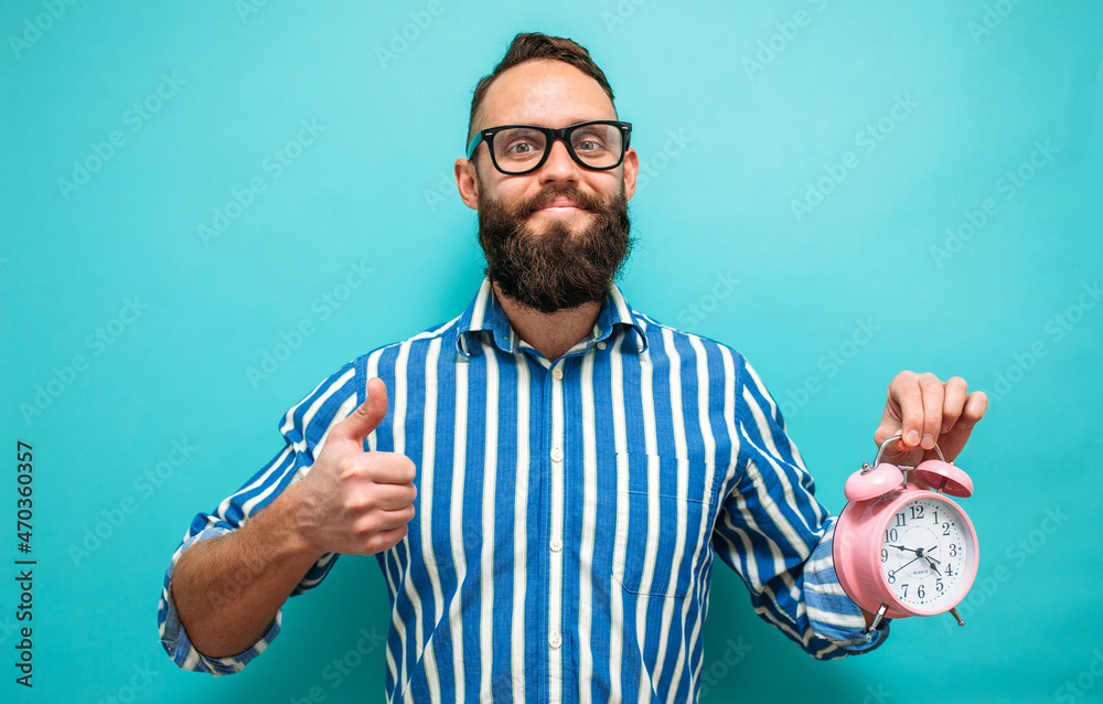 Young caucasian man with glasses and beard holding alarm clock on blue isolated background. Crazy emotions. The time has come - obrazy, fototapety, plakaty 