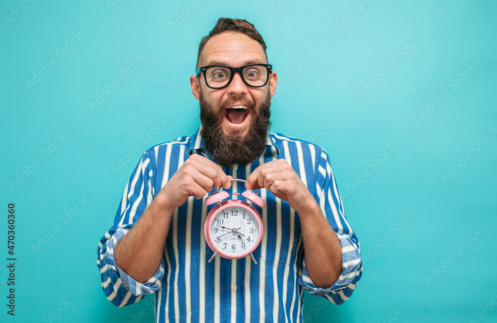 Young caucasian man with glasses and beard holding alarm clock on blue isolated background. Crazy emotions. The time has come - obrazy, fototapety, plakaty 