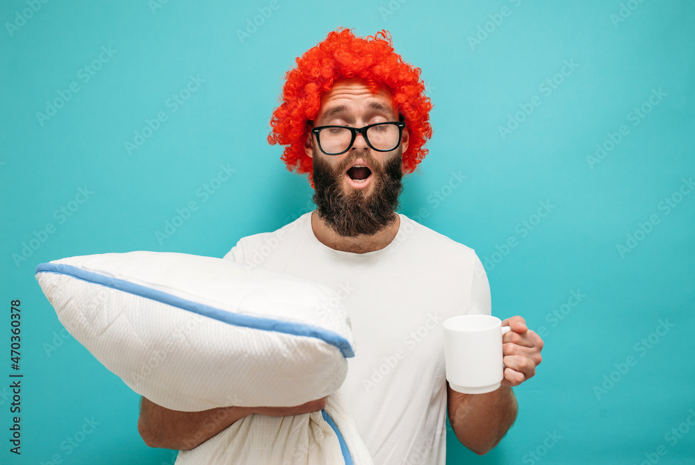 Morning awakening session. Stylish hipster guy with red hair in spectacles holds a pillow in his hands and a cup of coffee, his eyes closed and he yawns isolated on a blue background