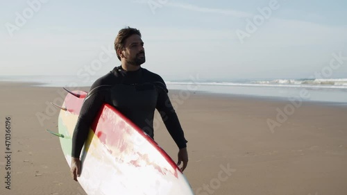 Medium shot of happy surfer with disability holding surfboard. Handsome man in wetsuit walking along sandy beach, looking for best place to enter ocean. Water sport, disability, surfing concept 