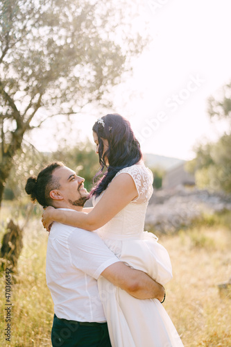 Groom holds bride in his arms against the background of trees on the lawn