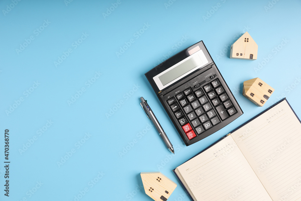 Top view desk office with the black calculator, little home models, and notebook on blue background with copy space. Concept loan, saving money for investment business property. Top view desk office with the black calculator, little home models, and notebook on blue background with copy space. Concept loan, saving money for investment business property.