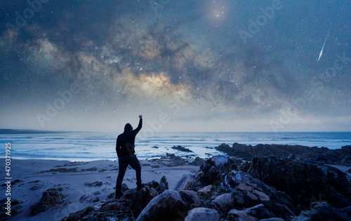 man standing on the rock  back view  with raised hand staring the Milky Way and stars over the ocean 