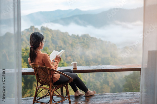 young woman reading book near window and looking mountain view at countryside homestay in the morning sunrise. SoloTravel, journey, trip and relaxing concept photo