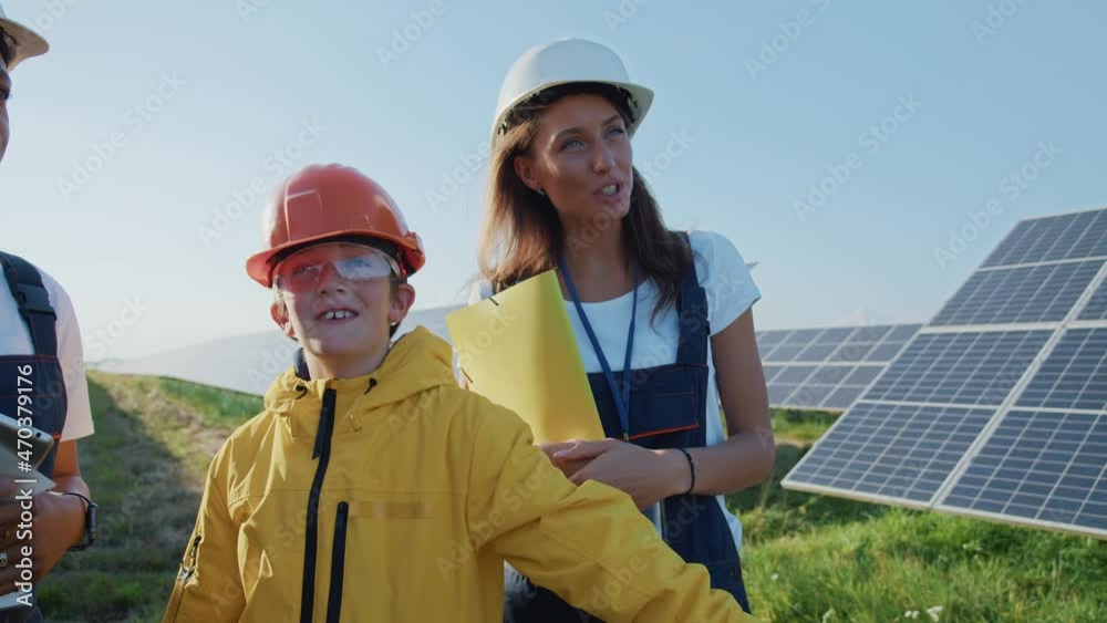 Cute teenager young boy wearing helmet talking with two female engineers walking on solar field. Scientific excursion. Alternative energy. Photovoltaic cells. Cute teenager young boy wearing helmet talking with two female engineers walking on solar field. Scientific excursion. Alternative energy. Photovoltaic cells.