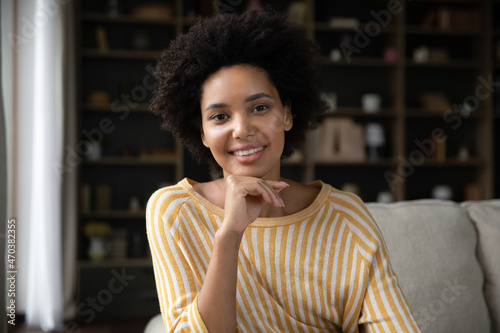 Happy millennial African American girl home head shot portrait. Smiling young woman looking at camera, talking on video call, sitting on couch, speaking online, touching face. Screen view